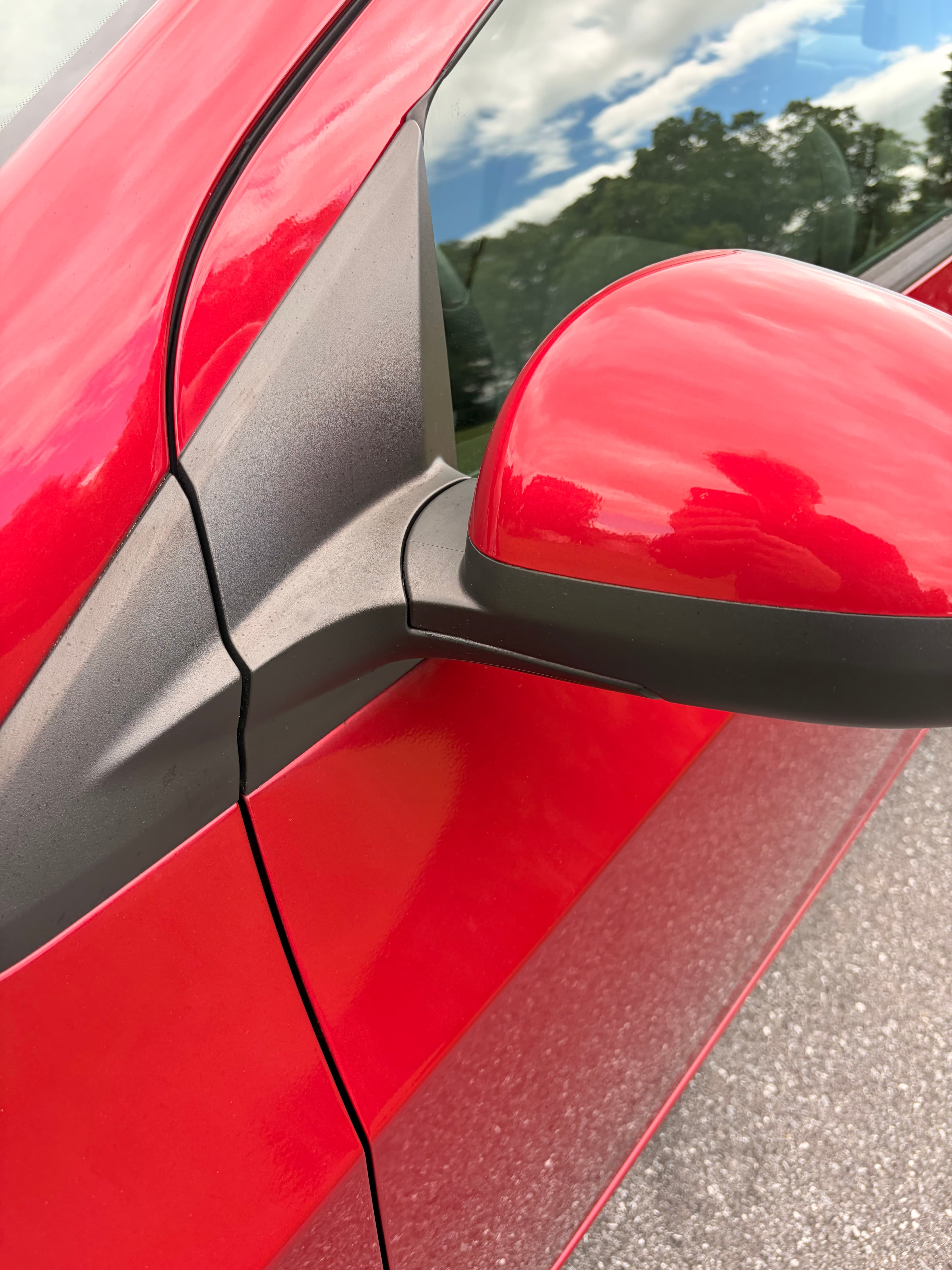 Bright red car side mirror and window reflecting a blue sky with white clouds.