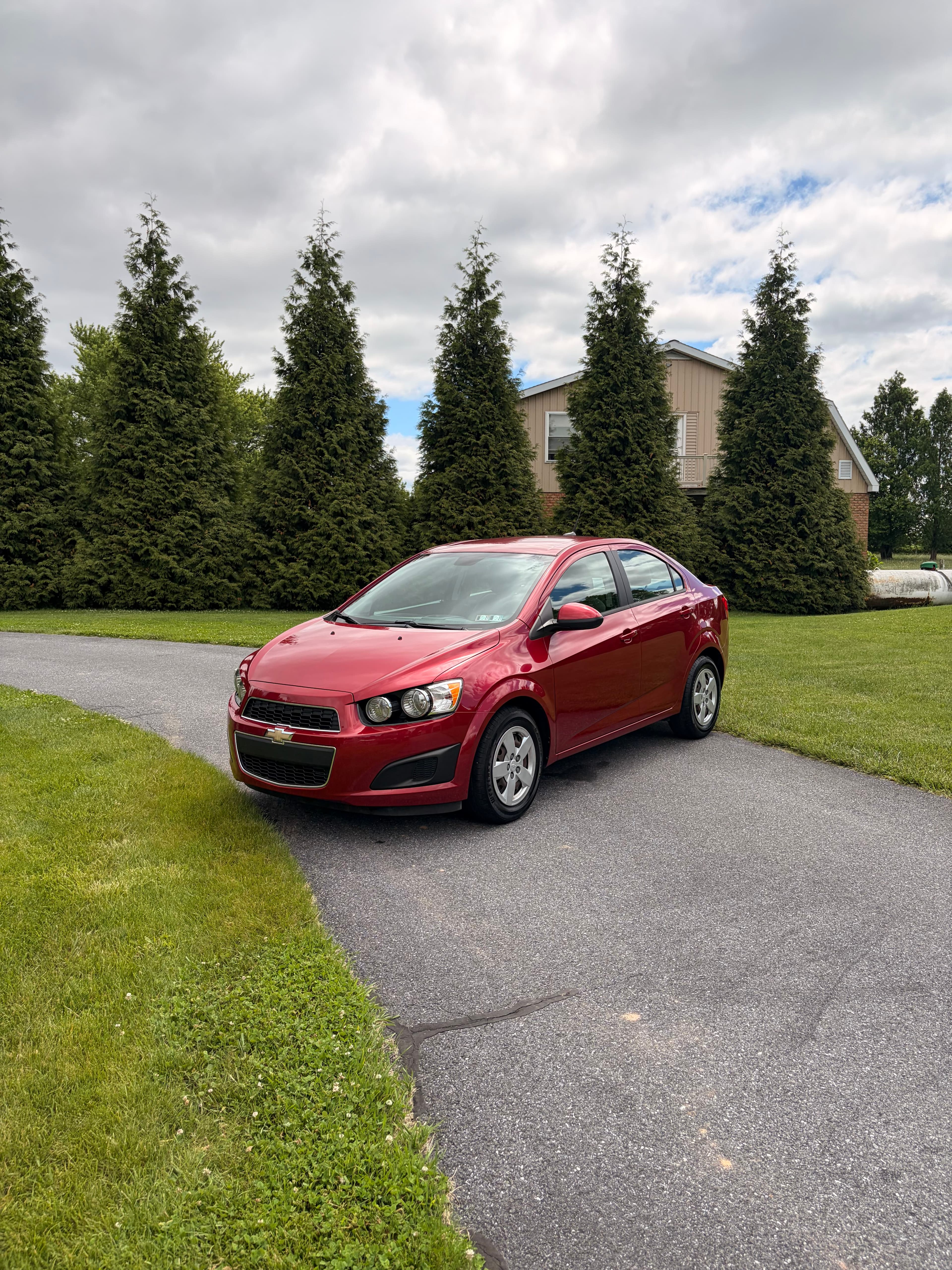 Red Chevrolet Sonic sedan parked on a paved driveway in front of tall evergreen trees.