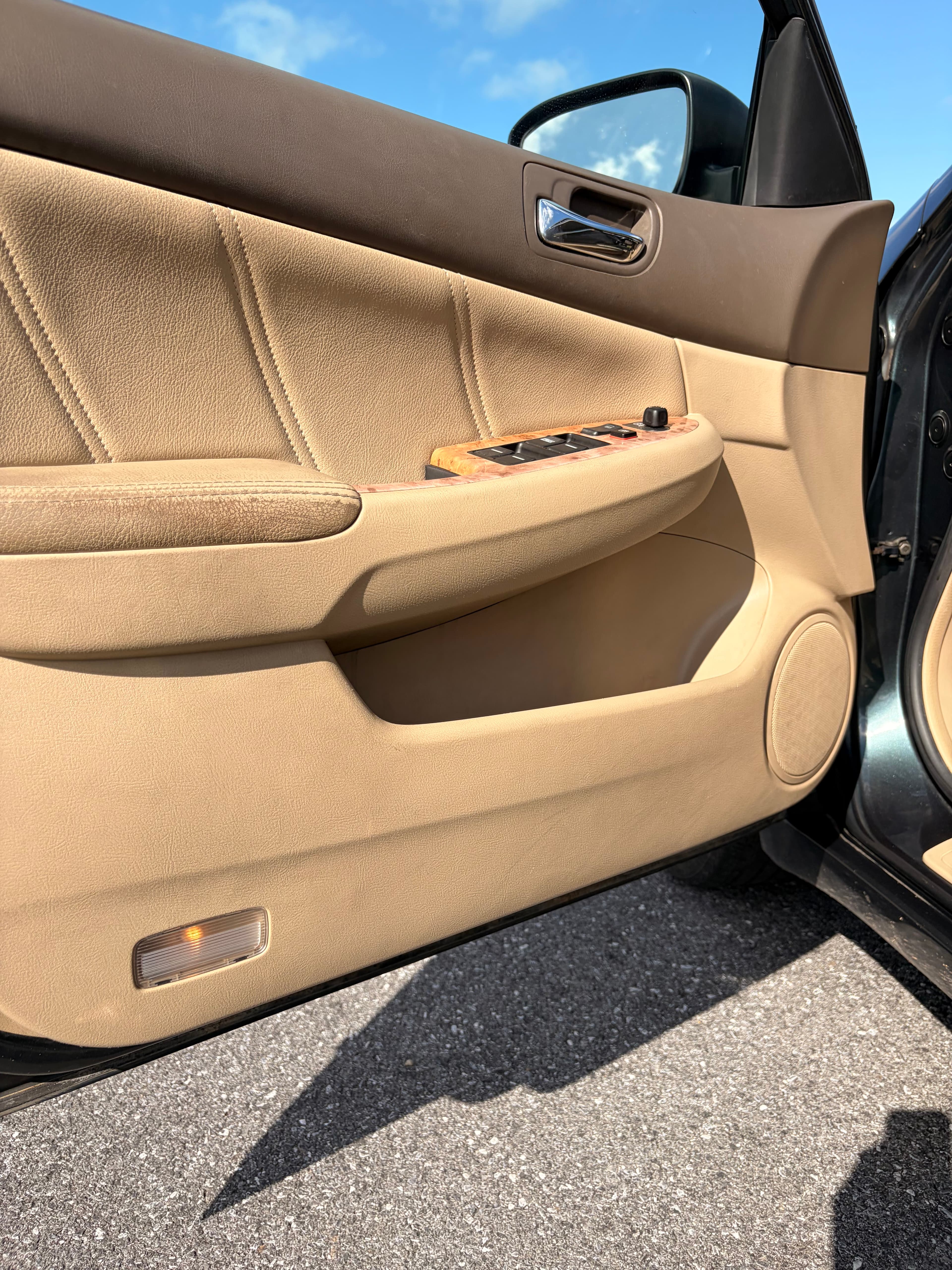 Open tan leather car door panel featuring wood-grain accents and window controls under sunlight.